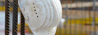 Ein weißer Schutzhelm hängt an einer Stahlarmierung auf einer Baustelle, umgeben von verschwommenen Baumaterialien und Gerüsten.