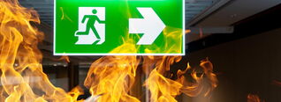 Ein grünes Notausgangsschild zeigt nach rechts, umgeben von Flammen. Es symbolisiert einen Fluchtweg in einer Notfallsituation mit Feuer im Gebäude.