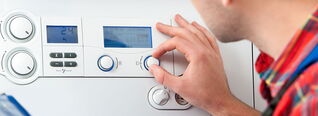 Eine Person dreht den Regler eines Heizkessels, um die Temperatur einzustellen. Im Hintergrund ist ein digitales Display mit Bedienelementen sichtbar.