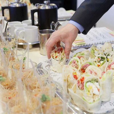 Hand greift nach Wraps auf einem Buffet mit Gläsern, Kaffeekannen und Tassen im Hintergrund