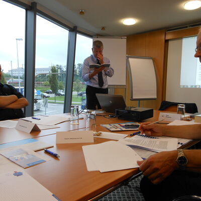 Drei Personen in einem Seminarraum mit Tischen, auf denen Papiere, Stifte und Namensschilder liegen, eine Person steht vor einem Flipchart und liest aus einem Buch