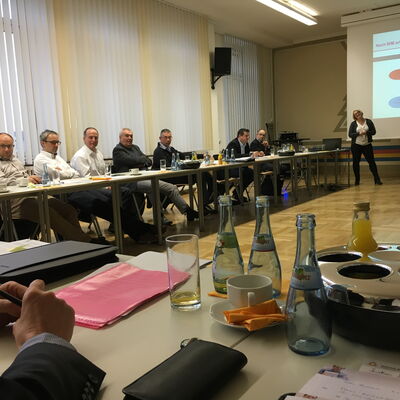 Mehrere Personen sitzen an einem langen Tisch in einem Seminarraum in Berlin, eine Person steht vor einer Projektionsfläche mit einer Präsentation.