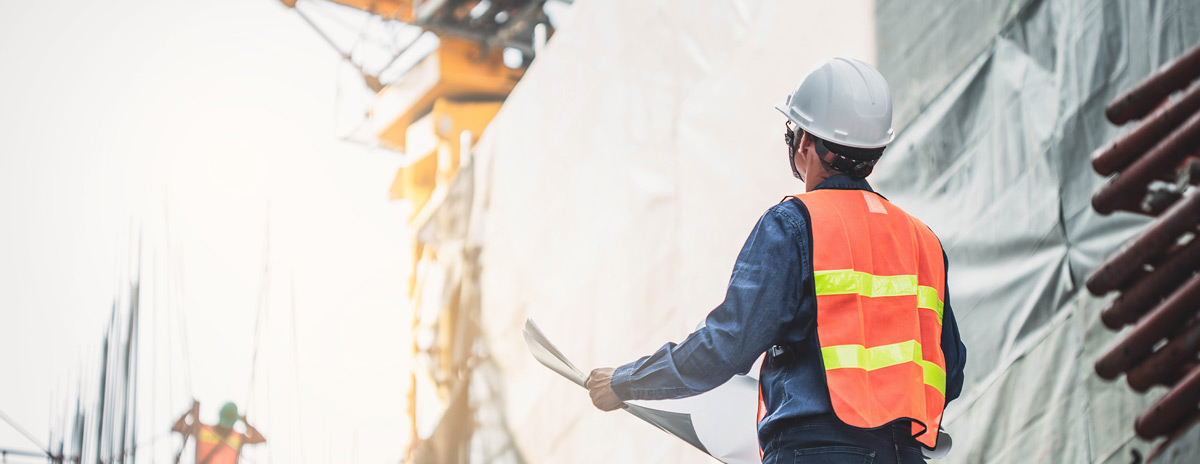 Ein Mann mit weißem Schutzhelm und roter Warnweste steht mit Plänen auf einer Baustelle und schaut zu einem Kran hoch.